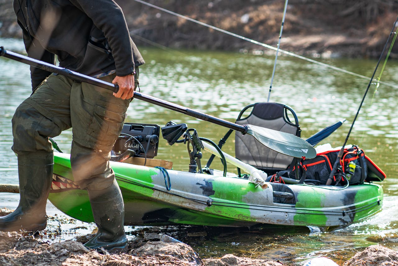 A man prepares a kayak on the riverbank for a fishing trip, holding a paddle.